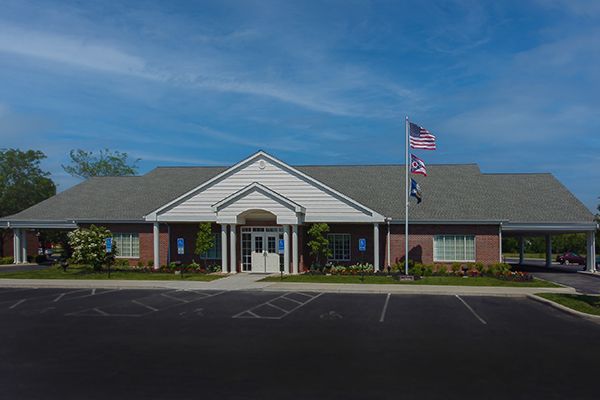 Exterior of a brick building with a white entrance, a flag pole, and a parking lot on a sunny day.