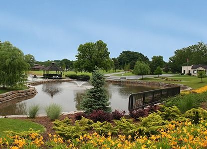 Lush park with a pond, gazebo, and flowers under a cloudy sky.