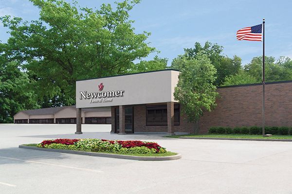Newcomer Funeral Home building with sign, American flag waving, red flowers, and trees.