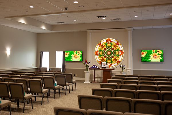 Interior of a chapel with rows of chairs, screens, and a stained-glass centerpiece on a table.