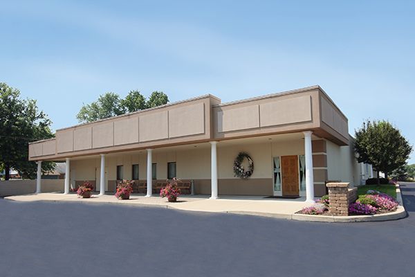 Exterior view of a one-story beige building with pillars, a wreath, and flower pots, likely a business.
