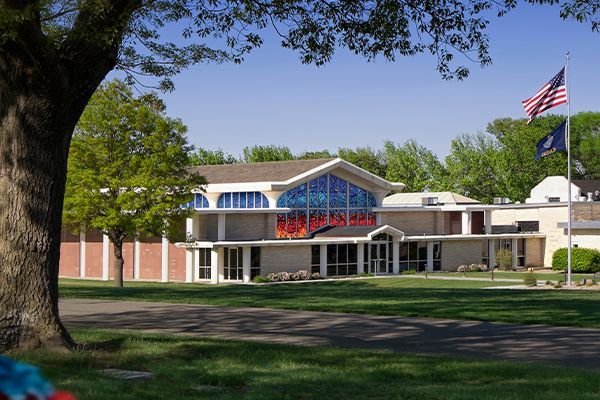 Church building with stained glass, flags, and a tree on a sunny day.
