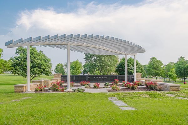 White pergola over a memorial with flowers, green grass, and trees under a cloudy sky.