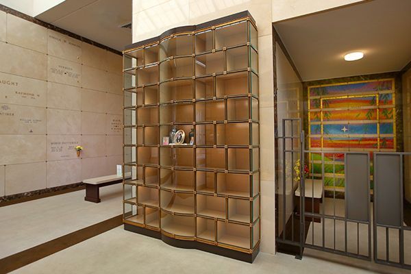 Interior of a columbarium with niches for urns, bench, and stained-glass art in background.