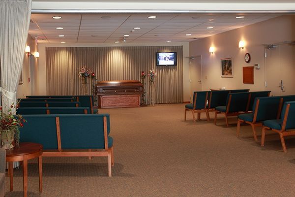 Funeral home chapel, brown casket centered, teal seating, beige carpet, a small TV screen on the wall.