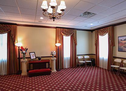 A funeral home interior. Red carpet, tan walls with brown trim, a chandelier, and chairs.