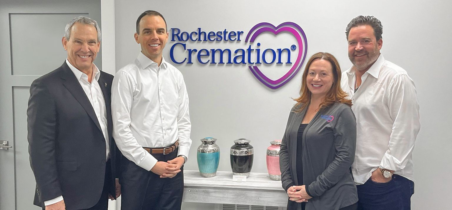 Four people stand in a funeral home. Rochester Cremation logo in the background. Three urns sit on a table.
