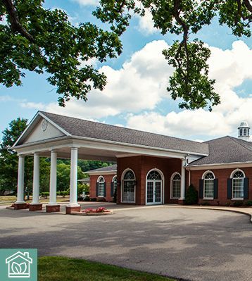 Red brick building with white columns, under a blue sky with fluffy clouds, surrounded by trees.