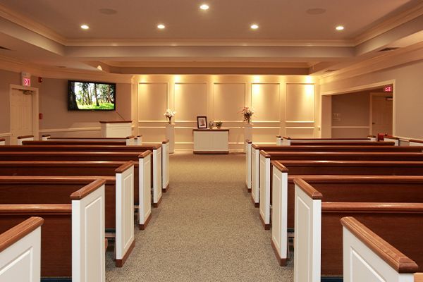 Rows of wooden pews in a funeral home chapel, leading to a central altar. Neutral tones, a screen.