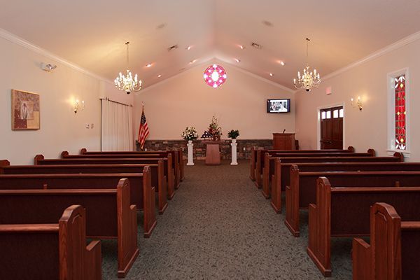 Interior of a chapel with rows of wooden pews facing a raised platform with a podium and floral arrangements.