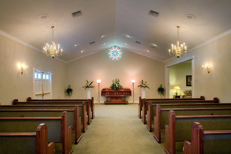 Interior of a funeral home chapel with rows of pews facing a casket and stained glass window.