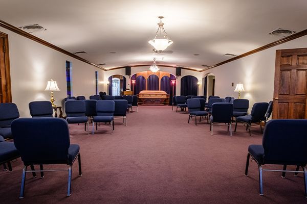 Funeral home chapel with rows of blue chairs, purple carpet, and casket at the front.