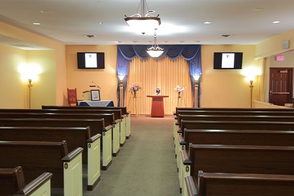 Interior of a funeral home with rows of pews, a stage, and floral arrangements.