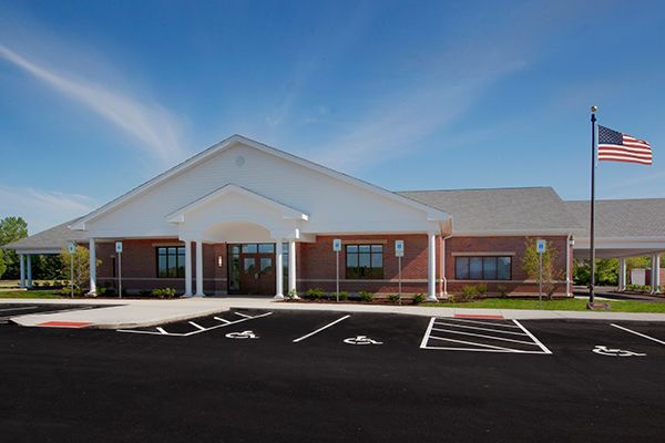 Brick building with a white roof and covered entrance, American flag flying, sunny sky, and parking lot.