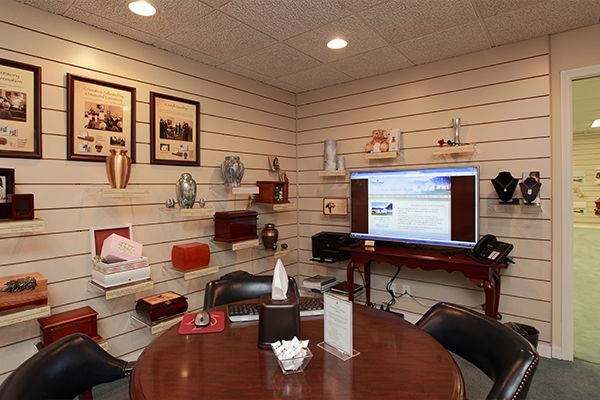 A funeral home sales room displays urns on shelves and a TV screen, with a table and chairs for consultations.
