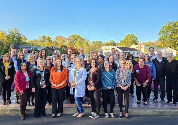 Group of diverse people standing outside, smiling. Autumn setting, trees in background.