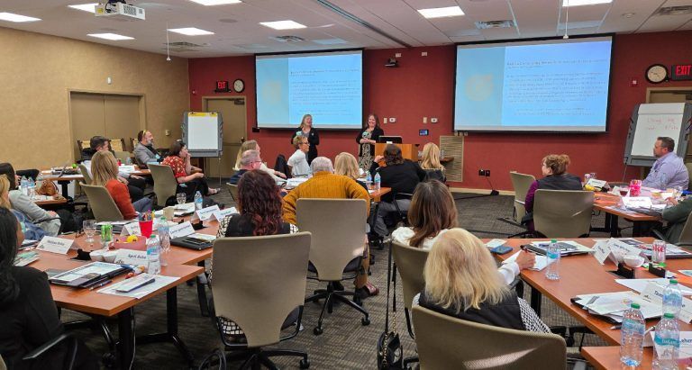 People attend a conference, with two presenters, two screens, and tables set up.