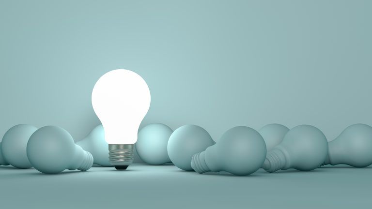 Glowing lightbulb stands out among many unlit bulbs on a light blue surface.