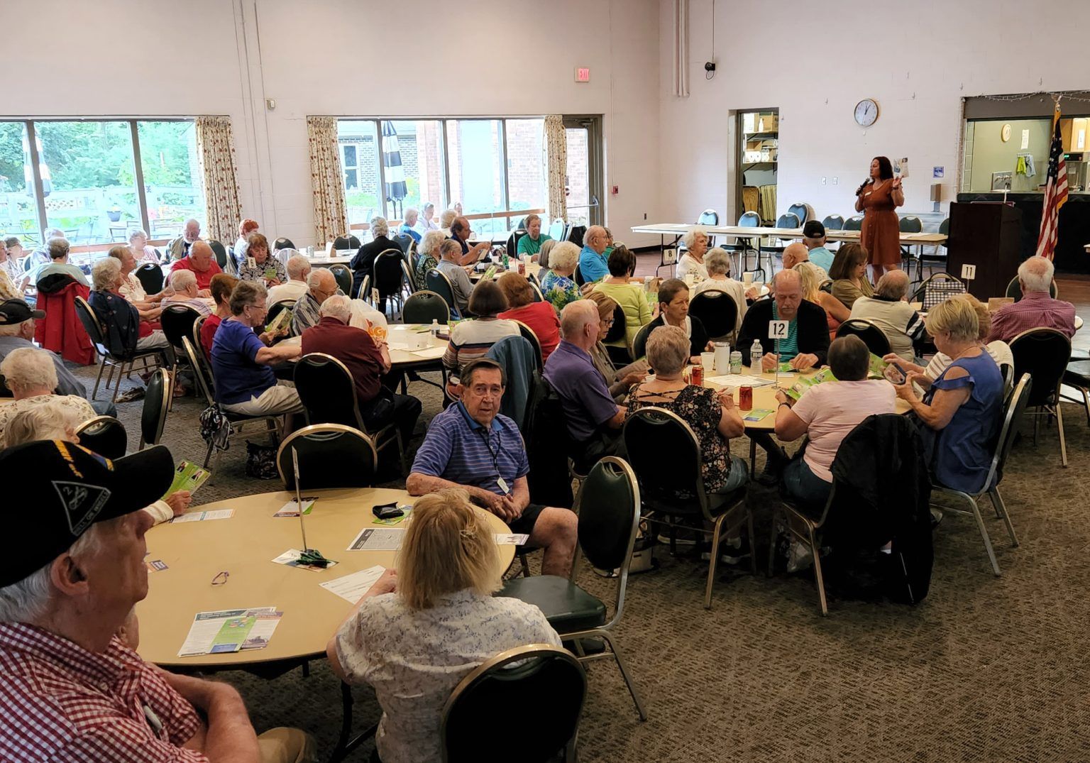 A large room with many seated people, some listening to a speaker. Daylight streams in.