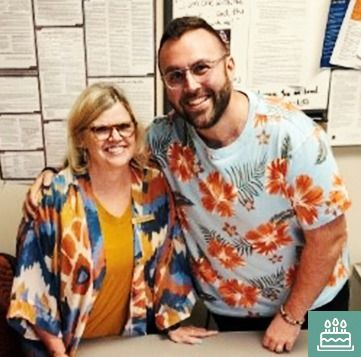 Woman with blonde hair and man with a beard smile together. Man wears a Hawaiian shirt, and they stand in an office.