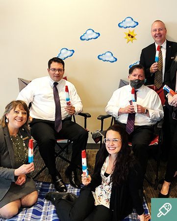 Group of people in office setting, holding popsicle treats. Decorated with paper sun and clouds.