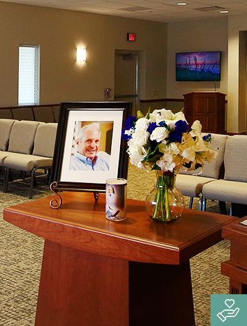 Memorial service: portrait of smiling man, flowers, and a candle on a wooden table, with rows of chairs in the background.