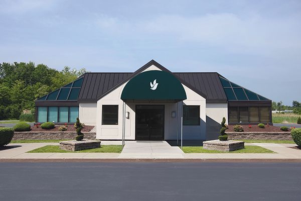 Off-white building with dark glass roof, green awning, and small decorative trees; sunny day.