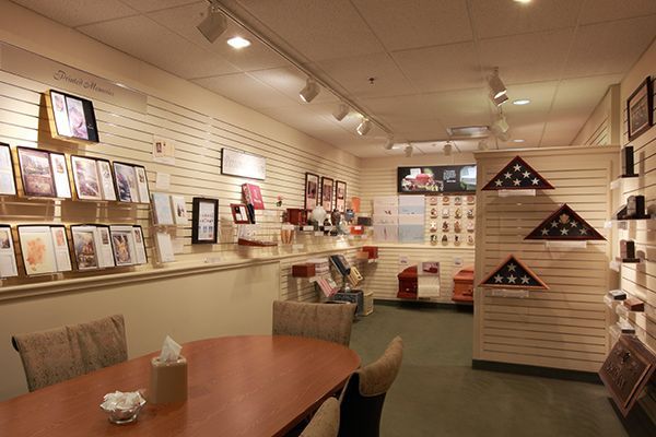Interior of a funeral home showroom with displays of urns, flags, and memorial items. A table with chairs is in the foreground.