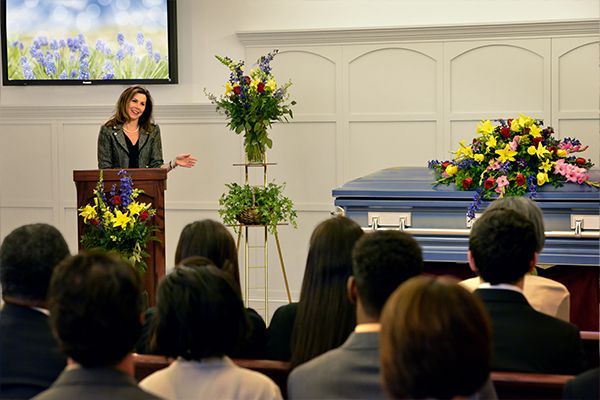 Woman speaking at a funeral, standing behind a podium. Attendees seated in pews. Blue coffin, floral arrangements present.