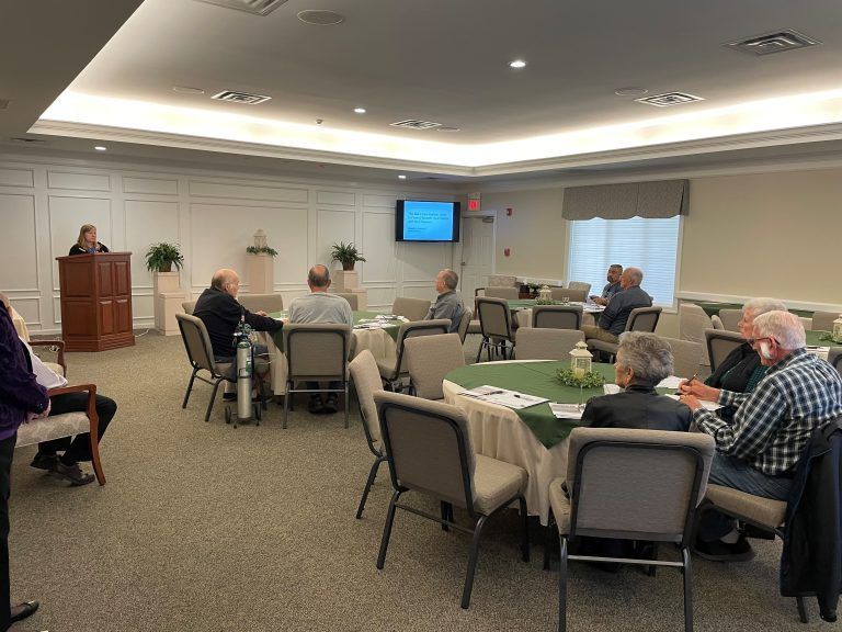 Meeting in a well-lit room: a speaker at a podium, attendees seated at round tables.