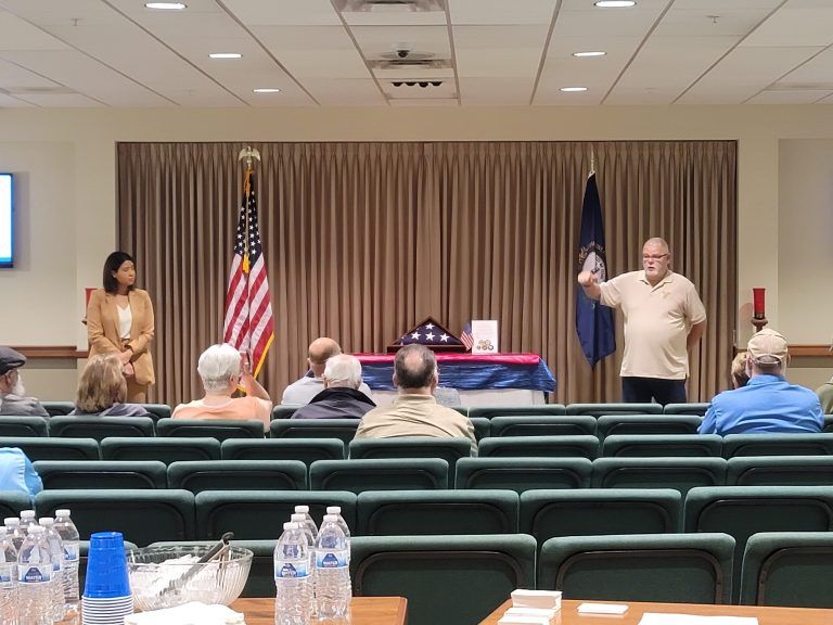 Man speaking at a presentation with an audience. Flags, memorial on a table. Indoors, neutral tones.