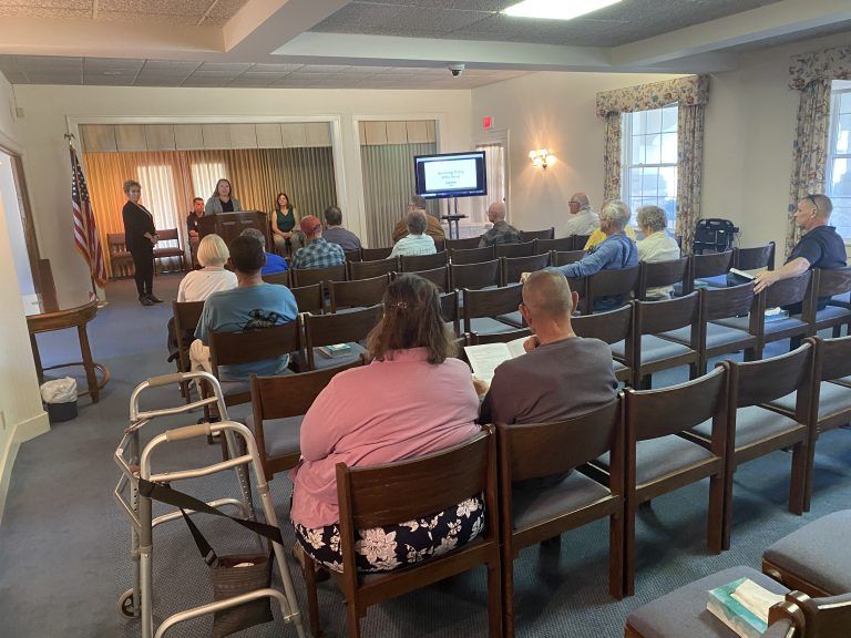 A presentation at a meeting. People seated, listening to presenters at the front. Room has windows, an American flag, and a TV screen.