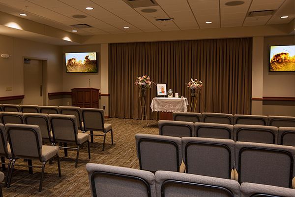 Funeral home chapel with rows of chairs facing a draped altar, two screens, and floral arrangements.