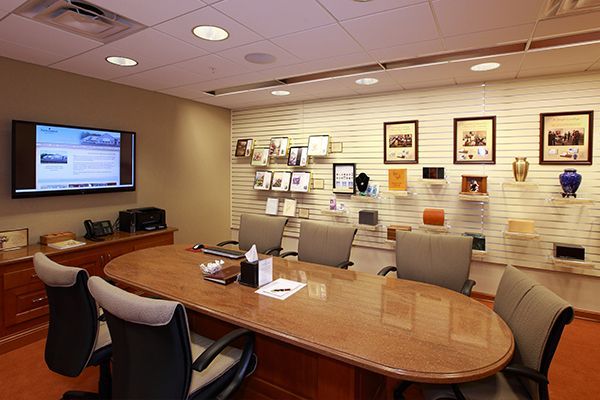 Meeting room with oval table, chairs, TV, and display of framed photos and urns on a striped wall.