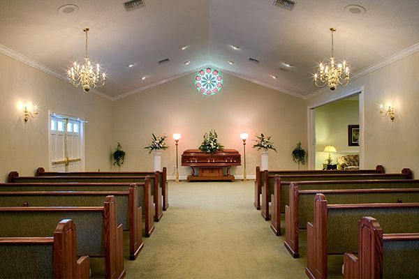 Interior of a funeral home chapel with rows of pews, a casket, and ornate chandeliers.