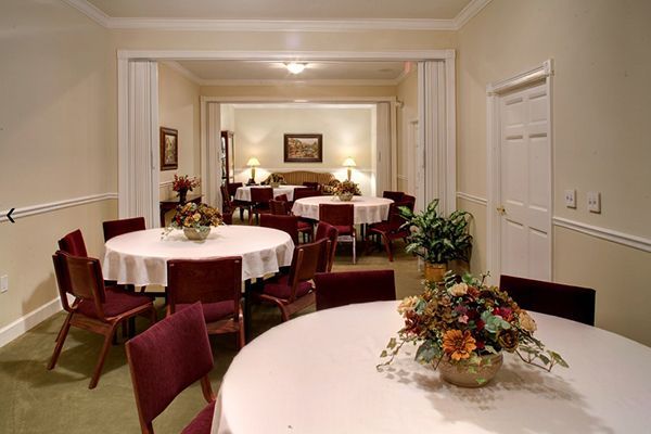 A dining room with round tables, chairs, and flower centerpieces. An open doorway leads to another room.