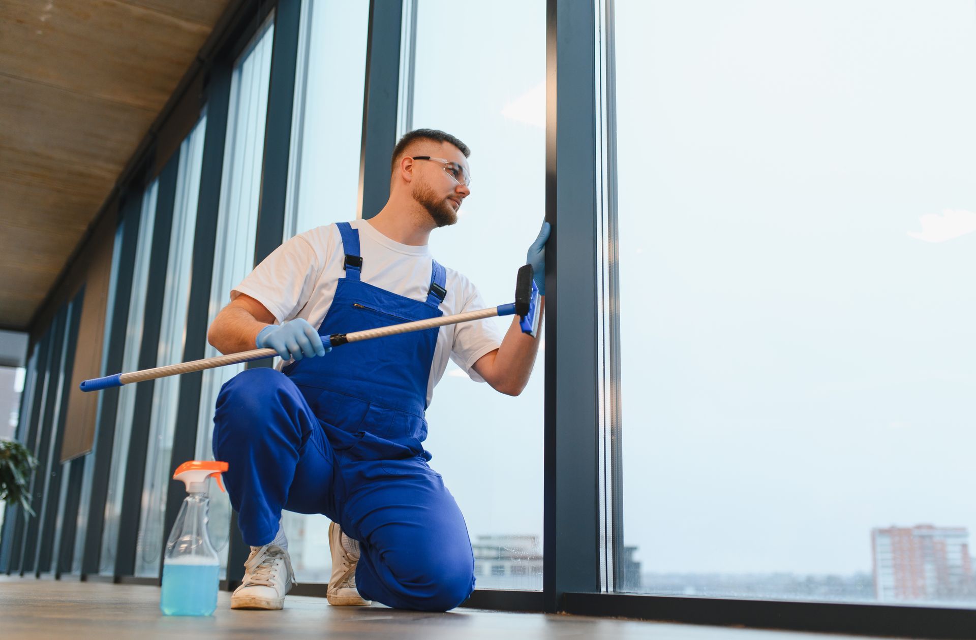 Worker in blue overalls cleaning large windows with a squeegee beside a spray bottle