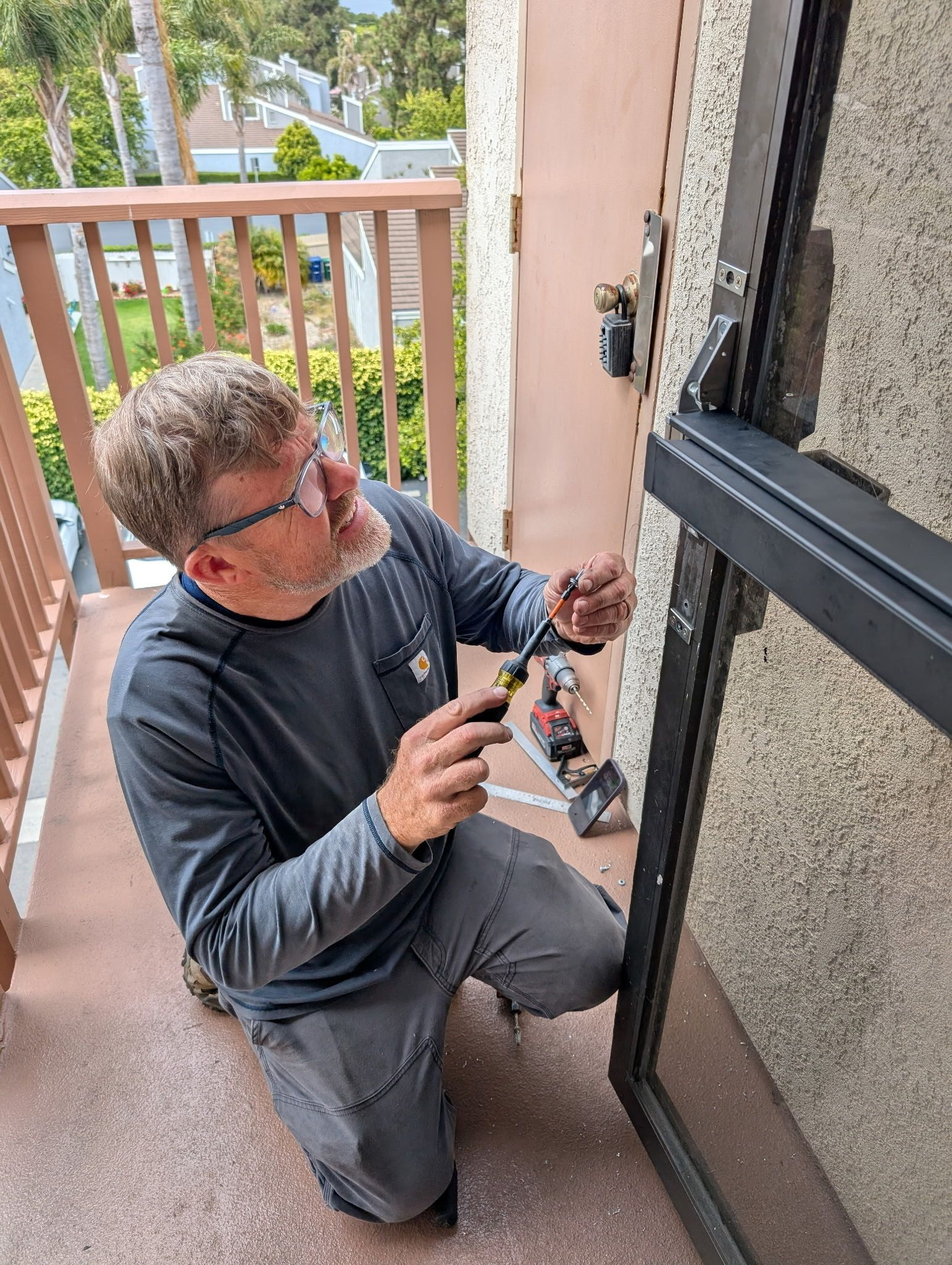 A man is kneeling down on a balcony working on a door lock.