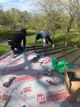 Two workers installing roofing underlayment on a sloped roof in a sunny orchard.