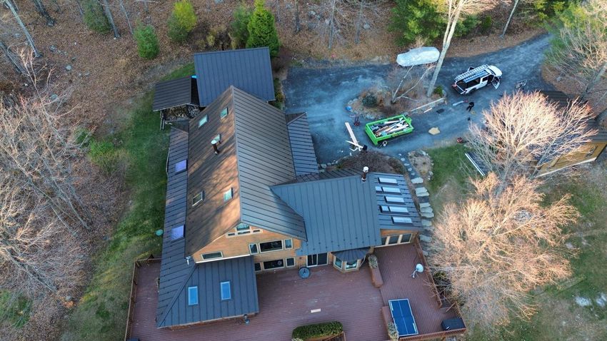 Aerial view of a dark-roofed house with a red brick patio, a green car, and a gravel driveway surrounded by trees.