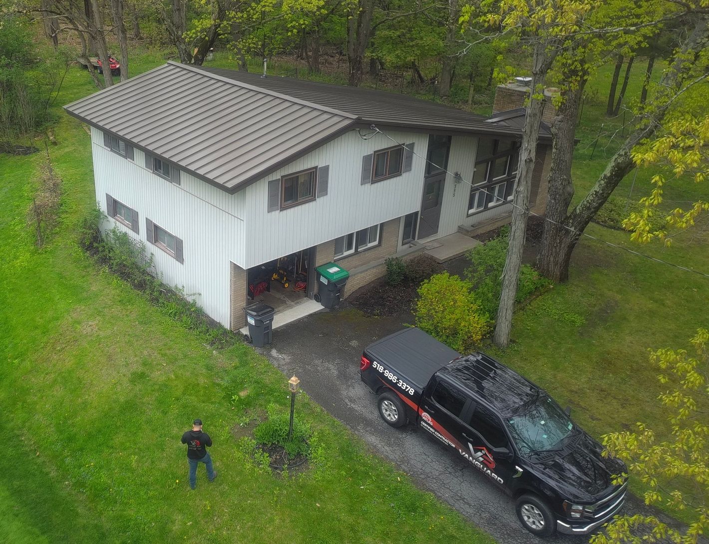 Aerial view of a white house in a wooded yard with a black pickup truck and one person outside.