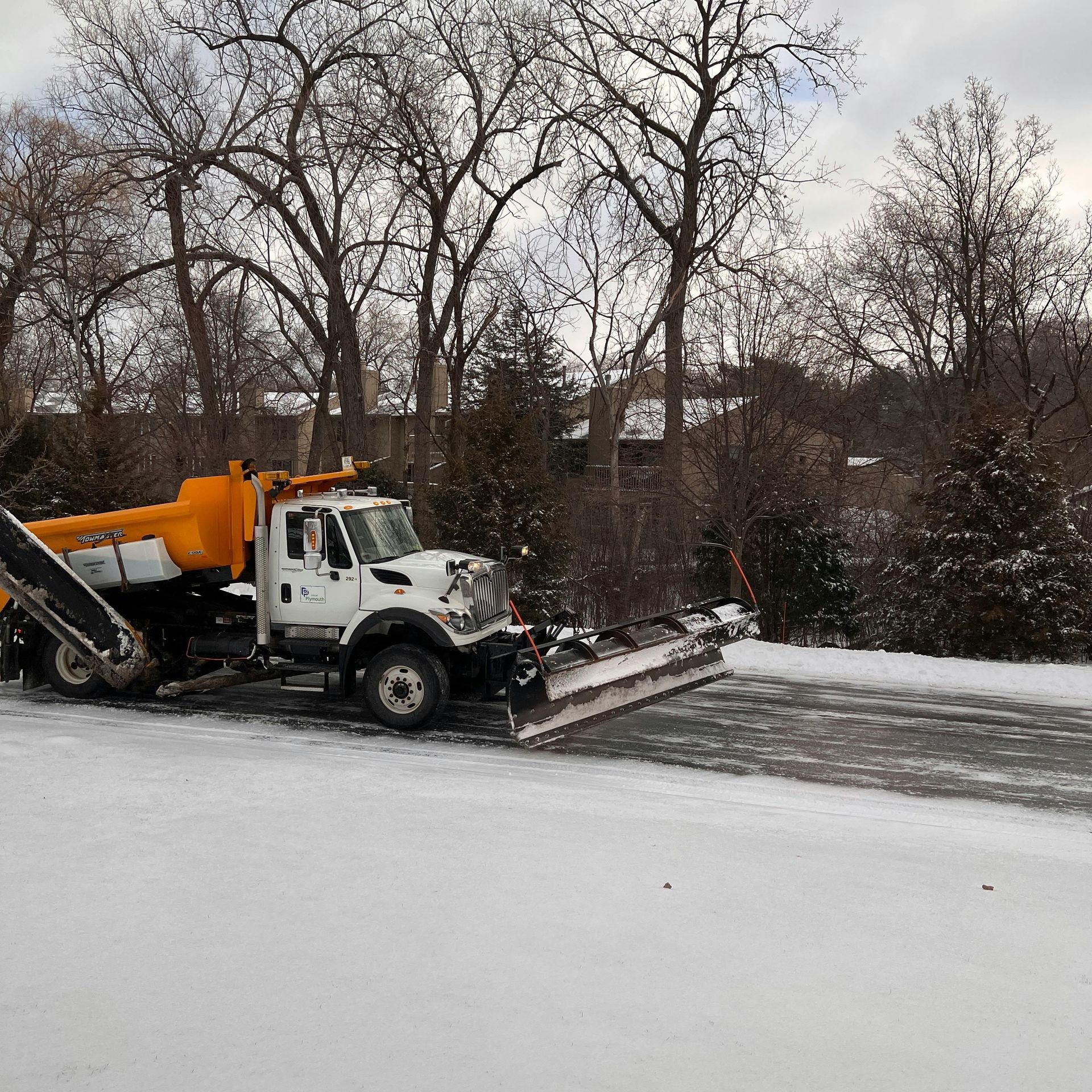 Snowplow clearing a residential street lined with snow-covered houses and trees on a cloudy day.