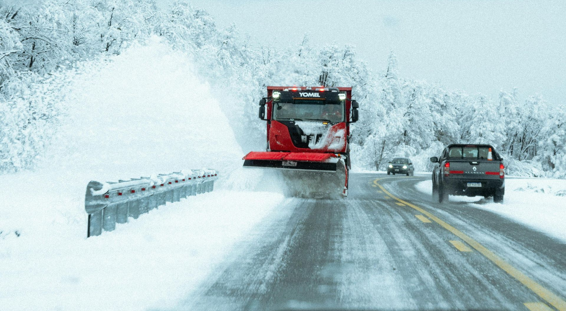 Snowplow clearing a snow-covered highway, with cars driving nearby, trees in the background, and snowy conditions.