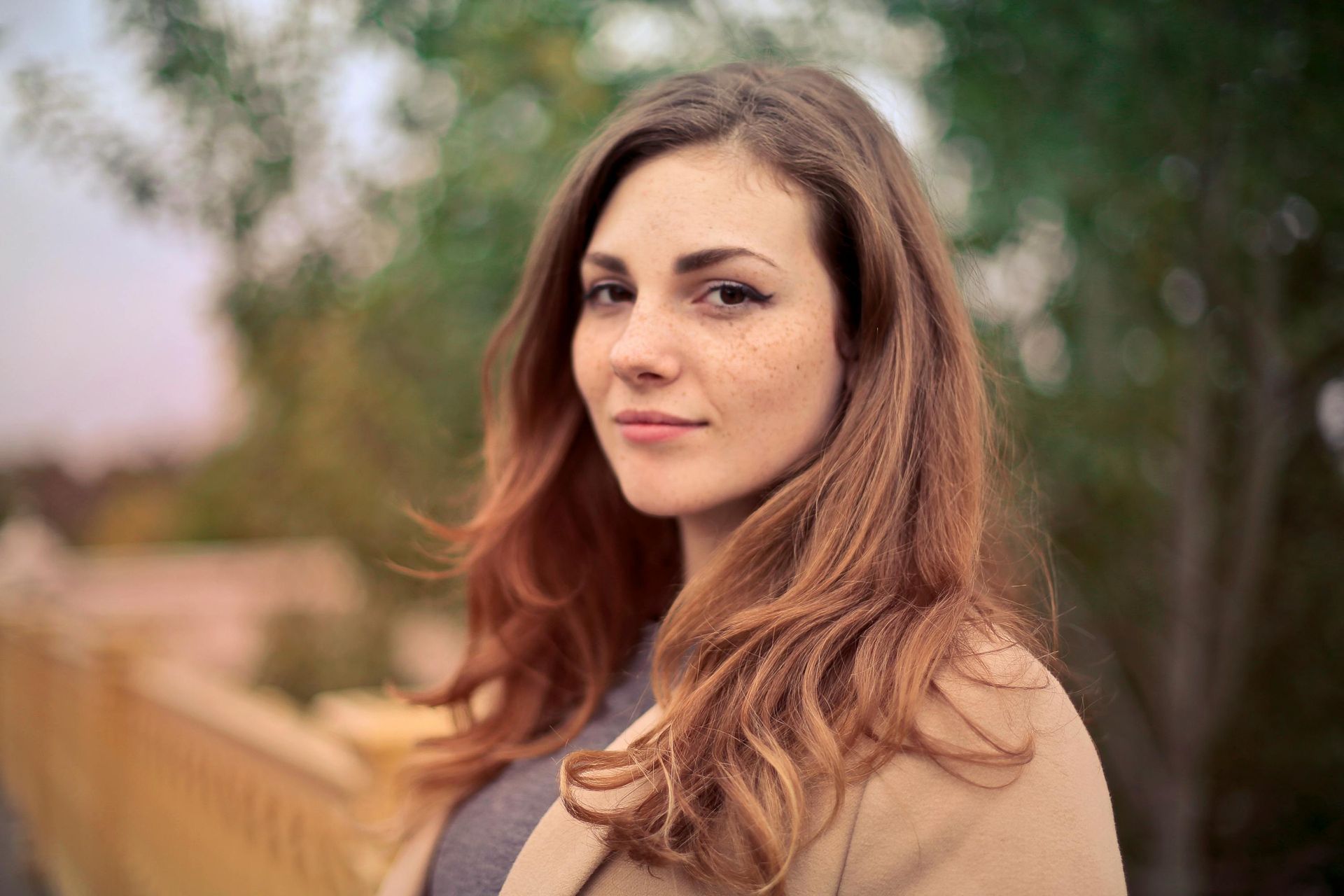 Woman with reddish-brown hair and freckles, wearing a beige coat, looks at the camera. Outdoor setting with greenery.