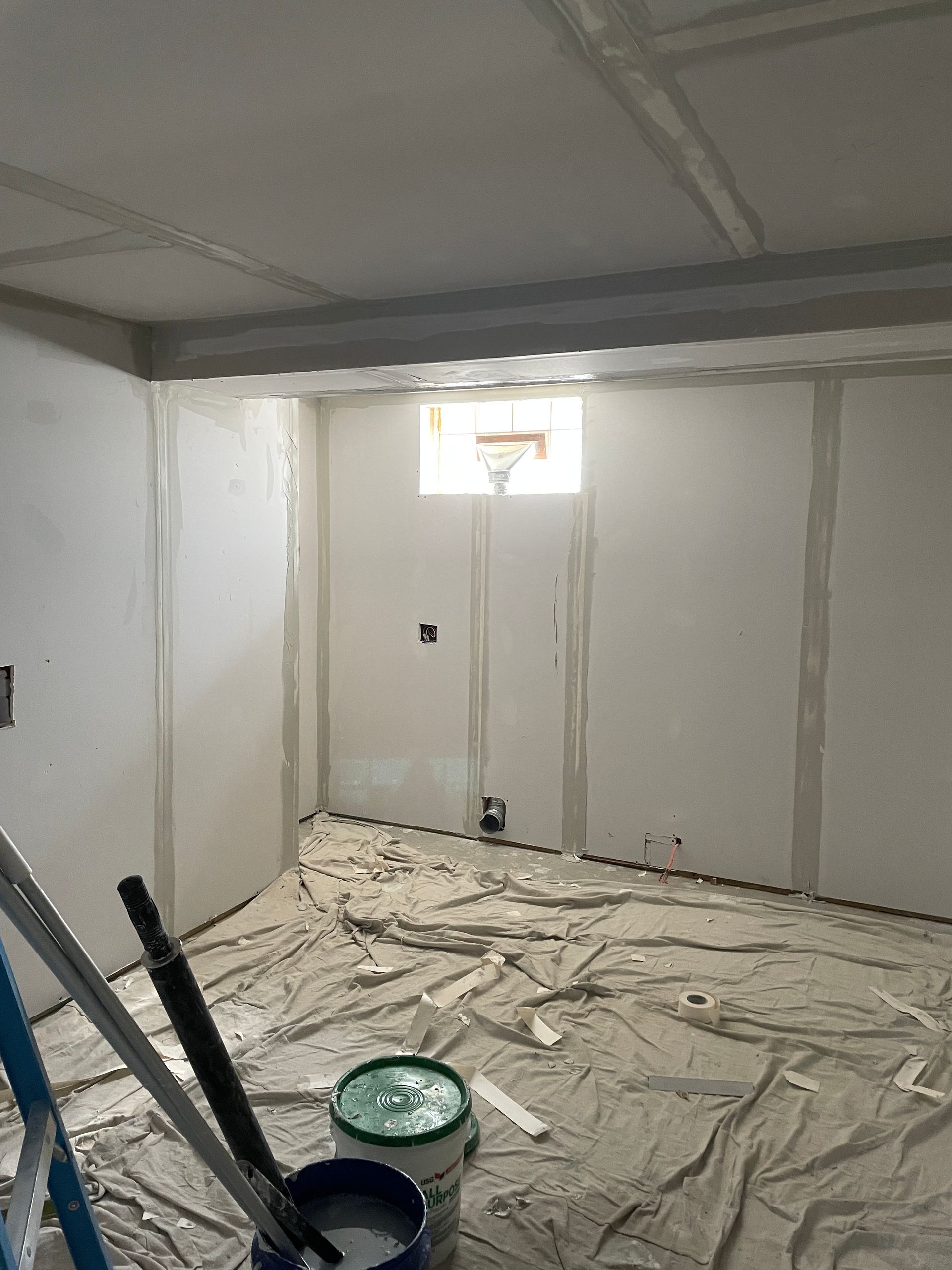 Room under construction with drywall, spackle, and protective covering on the floor. A small window is in the middle.