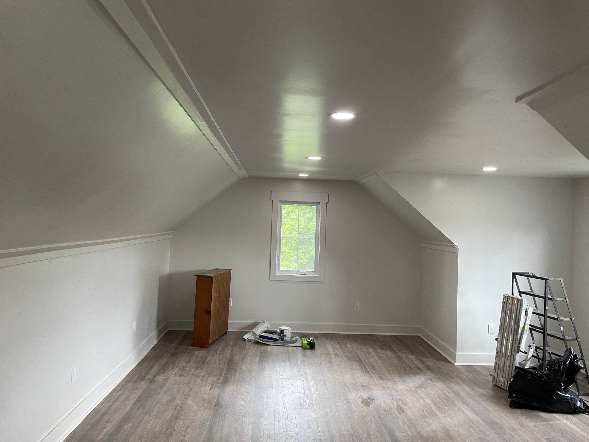 A room with white walls and ceiling, wooden floor, small window, and a wooden cabinet.
