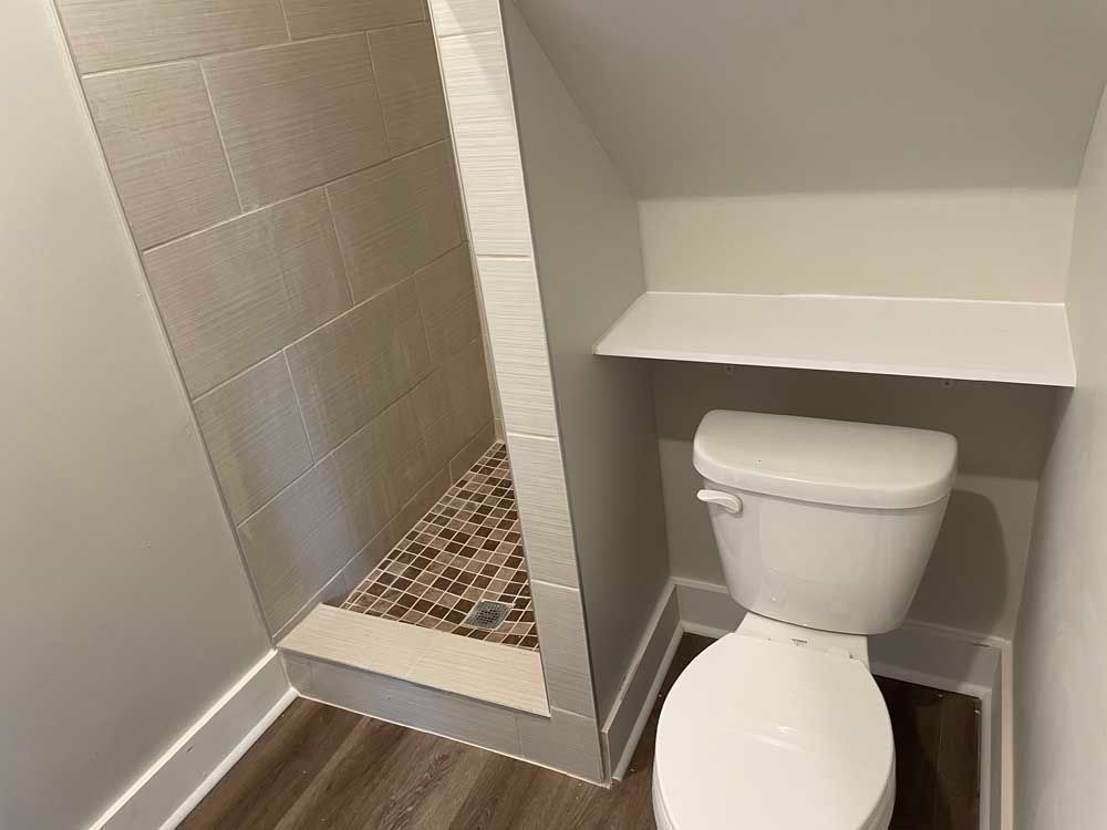 Bathroom with a toilet, shower, and shelf. The walls are light-colored, and the floor is brown.