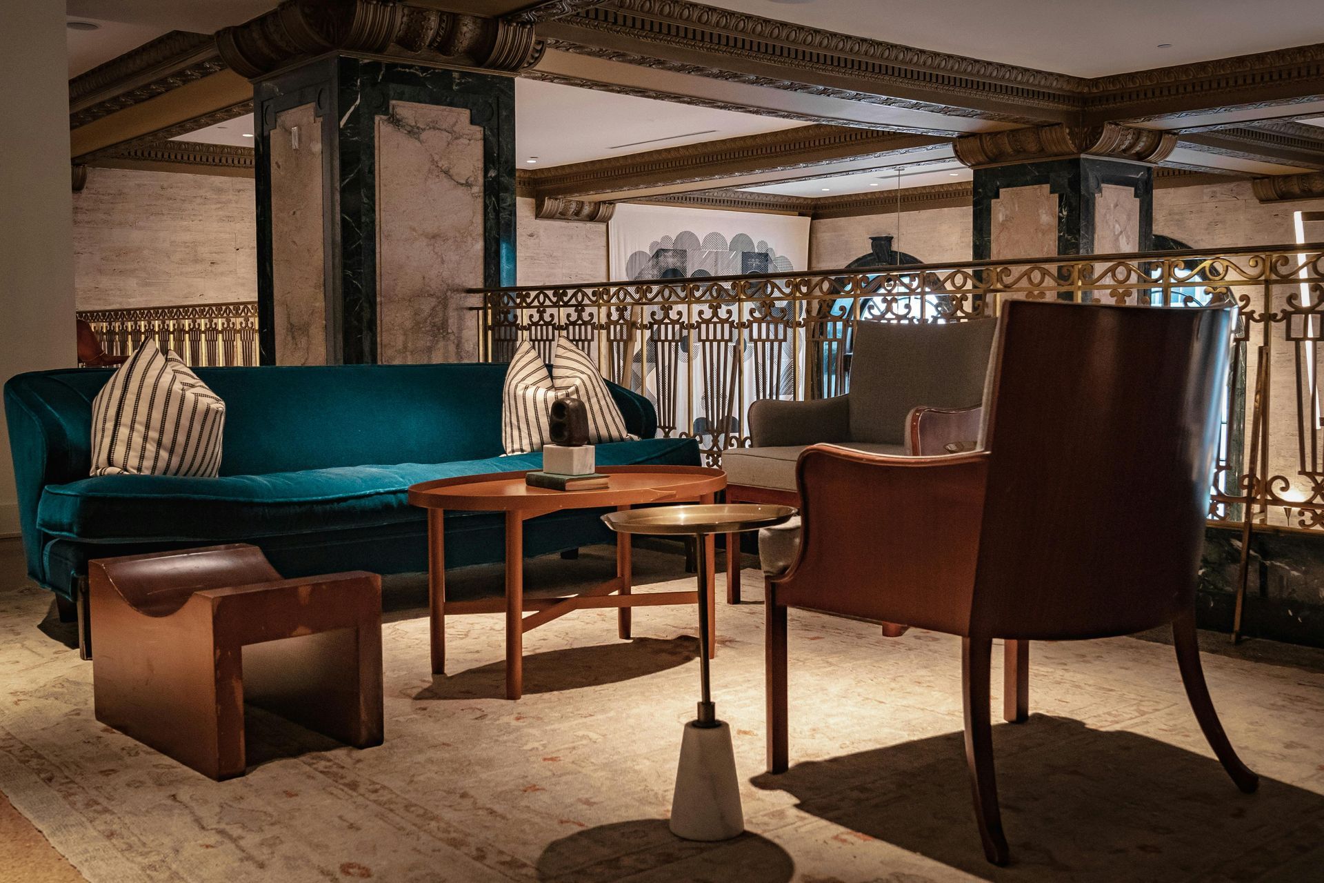 A teal sofa, a leather chair, and wooden tables arranged on a patterned rug in a room with ornate, classical ceiling beams.