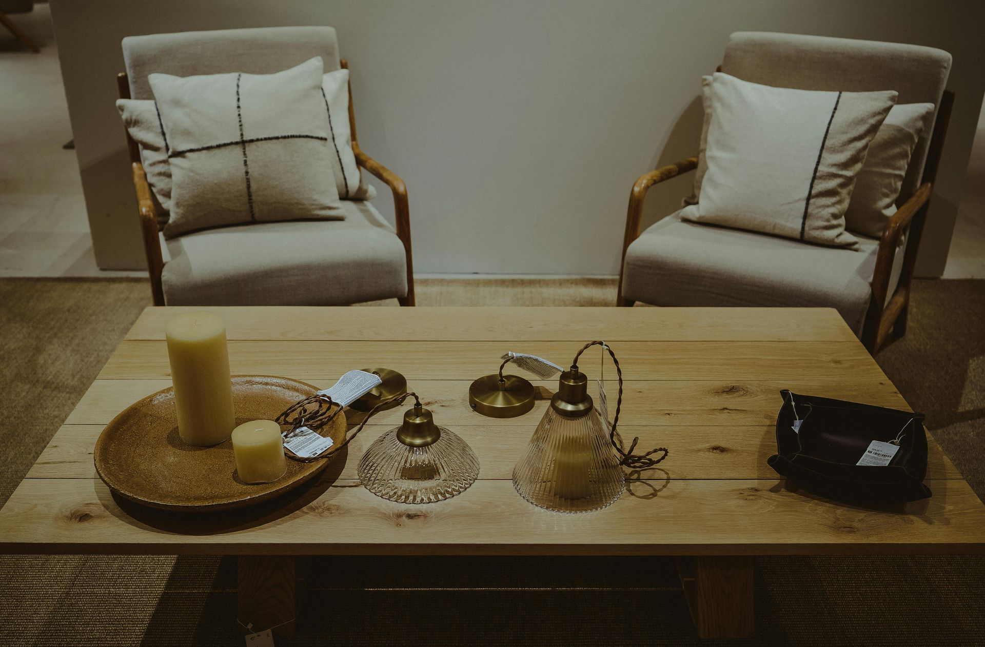 Two light-colored armchairs with decorative pillows face a low wooden coffee table holding candles and home accessories.