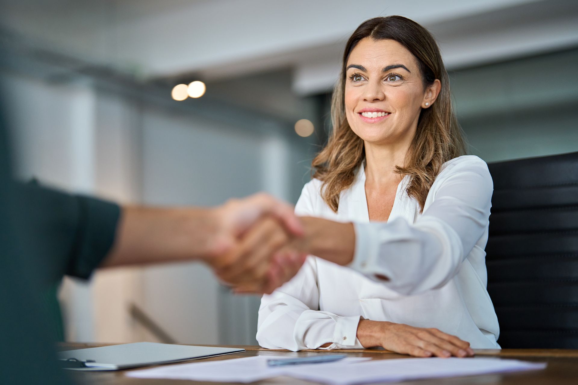 Woman in white shirt shaking hands with another person at a desk; smiling, indoors.